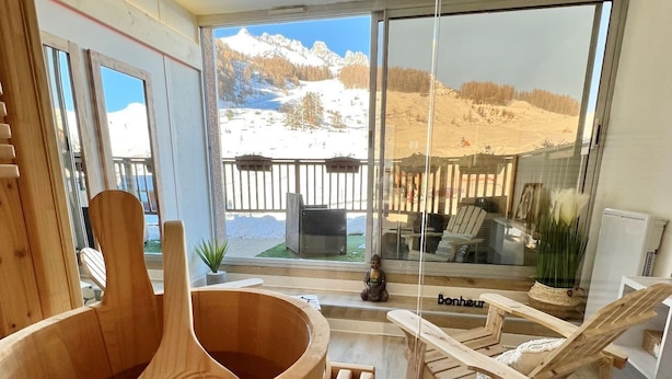 A wooden sauna with a large bowl and wooden benches, a wooden chair, and a view of a snowy mountain landscape through large windows. - Le Balcon Des Ecrins (Reallon)