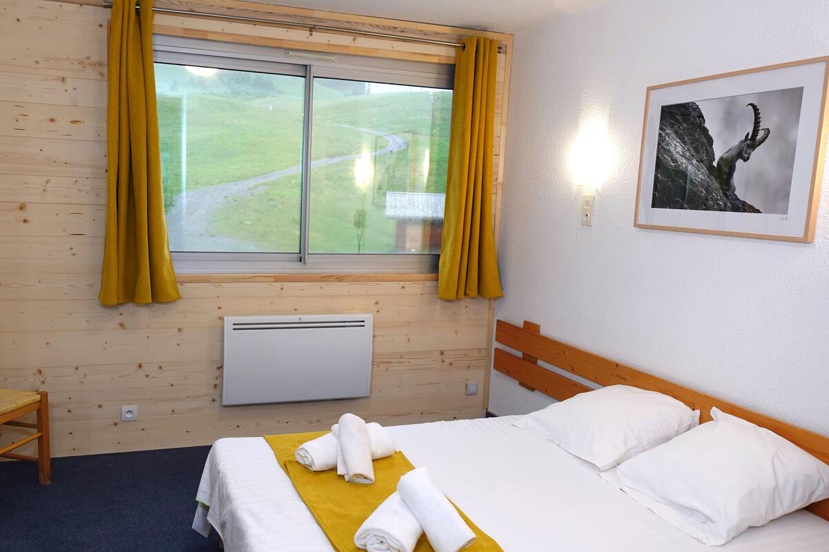 Ein Hotelzimmer mit Bett, Sessel, Fenster mit Vorhängen, Heizkörper und einem gerahmten Bild an der Wand.