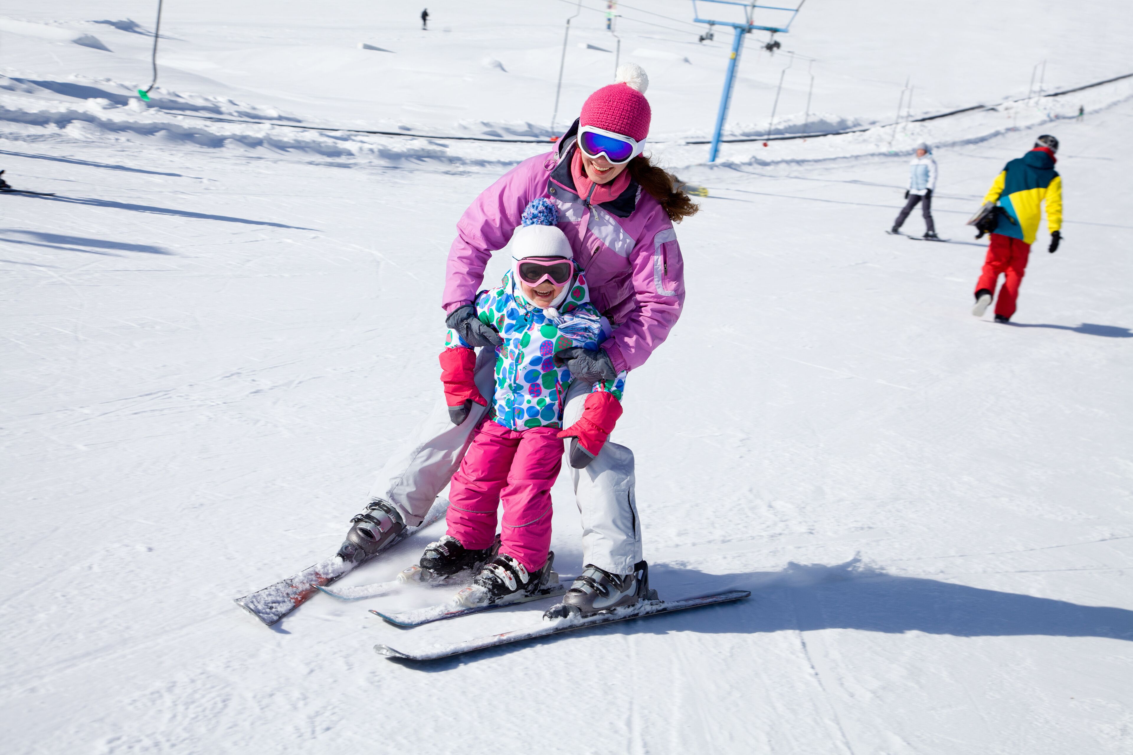 Ein verschneiter Skipisten mit Skifahrern und einem Kind, das von einer erwachsenen Person unterstützt wird.