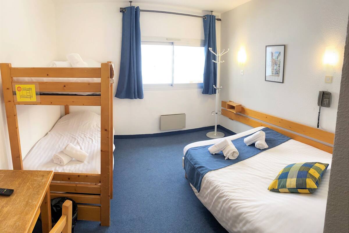 Ein Hotelzimmer mit Etagenbett, einem Einzelbett, einem Schreibtisch mit Stuhl, einem Kleiderständer, einem Fenster mit Vorhängen, einem an der Wand montierten Telefon und einem gerahmten Bild.