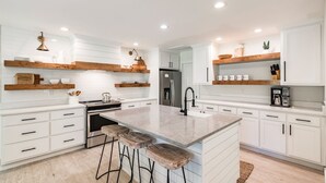Una cucina moderna con mobili bianchi, mensole in legno, un'isola centrale con sgabelli e un frigorifero in acciaio inossidabile.