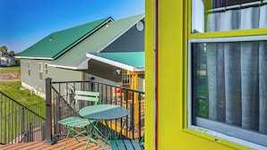 Uma varanda com uma mesa e cadeiras verdes, guarda-corpo amarelo e vista para uma casa com telhado verde.