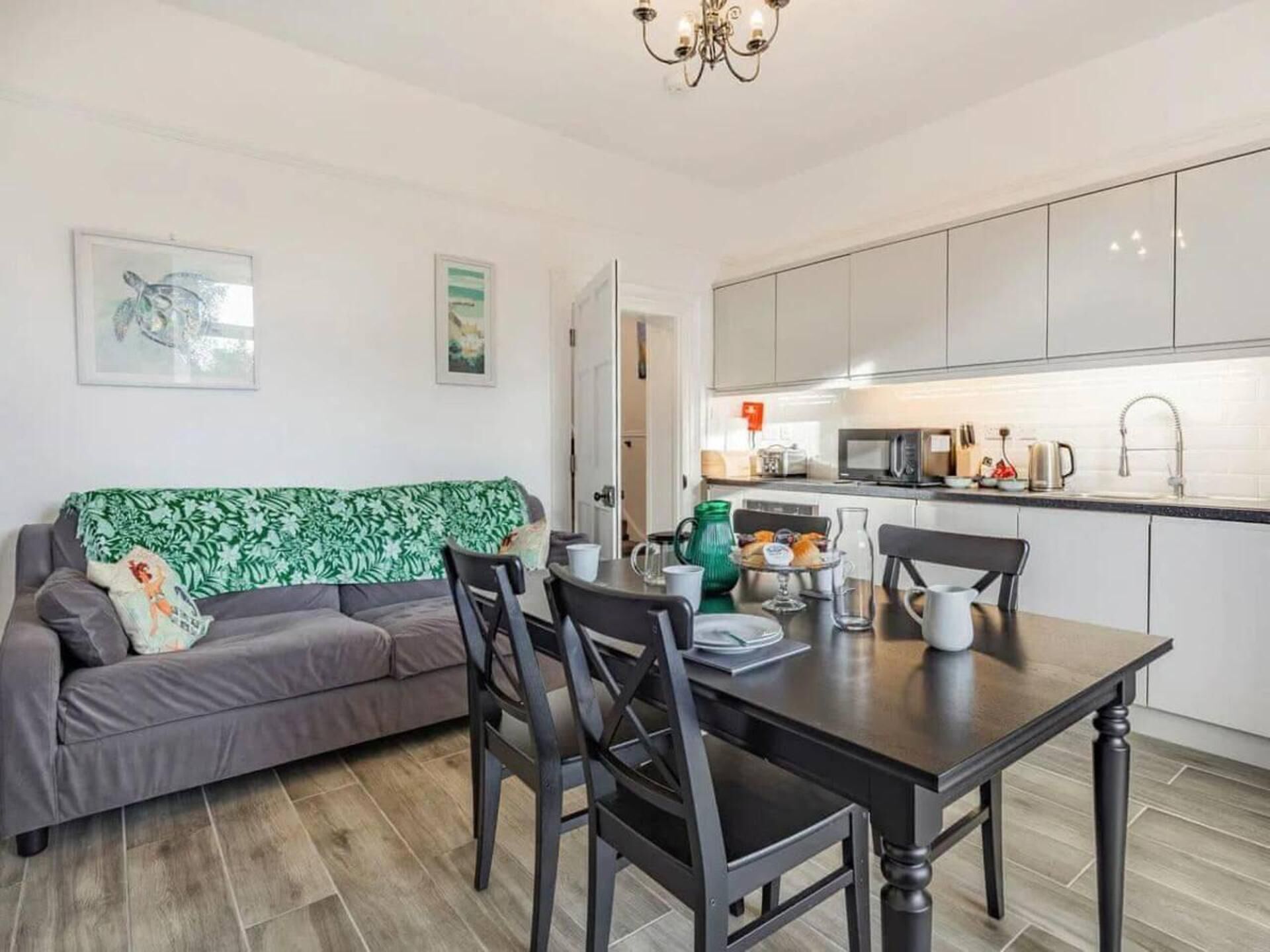 A modern living room with a grey sofa, a dining table with chairs, and a kitchen area with white cabinets and appliances.