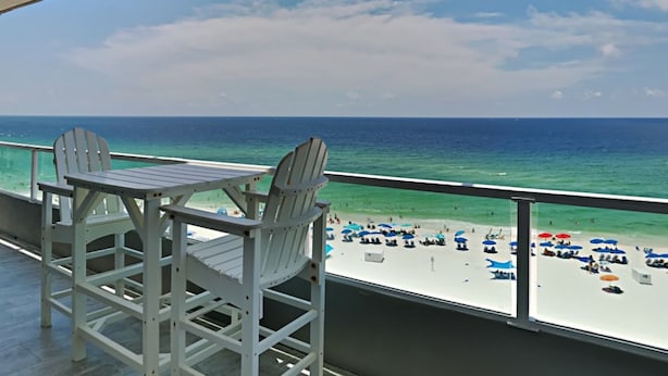 Un balcon avec deux fauteuils Adirondack donnant sur une plage parsemée de parasols et fréquentée par des gens.