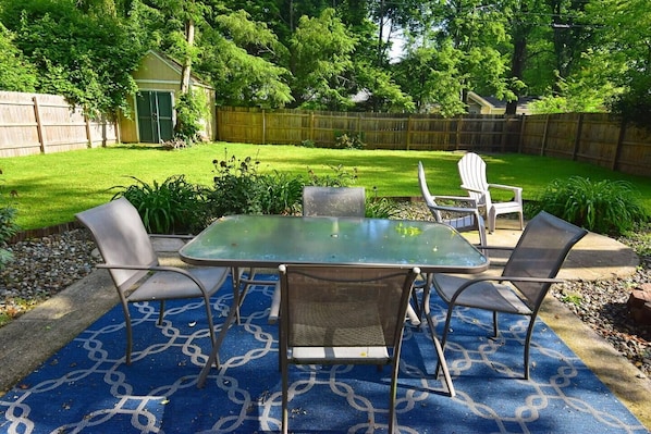 An outdoor patio with a glass-top table and four chairs, surrounded by a blue patterned rug.