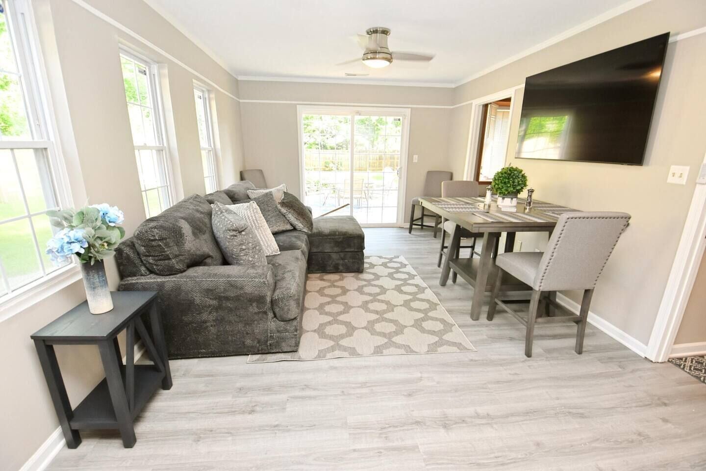 A modern living room with a grey sofa, a dining table with chairs, a TV mounted on the wall, and a large window providing natural light.