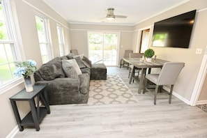 A modern living room with a grey sofa, a dining table with chairs, a TV mounted on the wall, and a large window providing natural light.