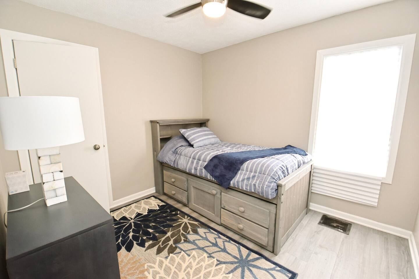 A bedroom with a bed, a desk, a lamp, a window, and a ceiling fan.