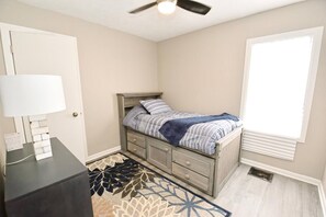 A bedroom with a bed, a desk, a lamp, a window, and a ceiling fan.