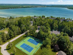 An aerial view of a lakeside property with two tennis courts surrounded by dense forest and residential buildings. - *NEW* Lakeside Club - 10 Minutes to Harbor Springs and Petoskey! (Petoskey)
