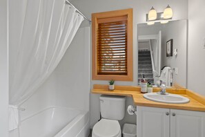 Et badeværelse med badekar, toilet, håndvask og et vindue med persienner.