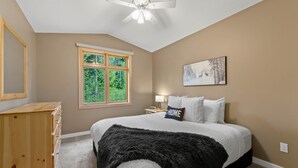Ein Schlafzimmer mit einem Bett, einem Deckenventilator, einem Fenster mit Blick auf Bäume, einer hölzernen Kommode und einem Bild an der Wand.