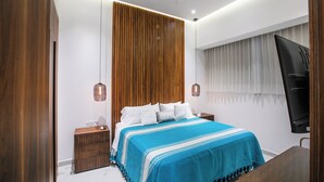 A modern bedroom with a wooden headboard, a bed with white bedding, a blue accent throw, and a TV mounted on the wall. - NEW Chic Condo with Scenic Views | Pool & Gym (Puerto Vallarta)