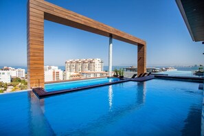 Une piscine à débordement située sur le toit, avec des poutres en bois, offrant une vue dégagée sur le paysage urbain et une mer calme en arrière-plan.
