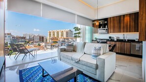 A modern living room with a sofa, coffee table, and chairs, a kitchen area, and a large balcony with a city view. - NEW Chic Condo with Scenic Views | Pool & Gym (Puerto Vallarta)