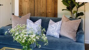 Ett vardagsrum med en blå soffa, dekorativa kuddar, ett glasbord med en bok och blommor, en träpanel på väggen och en hängande lampa.