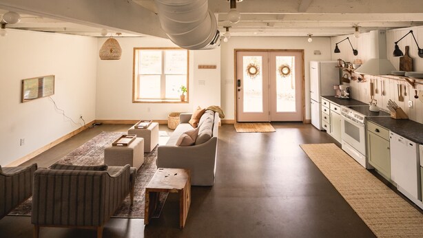 Een ruime woonkamer met een bank, stoelen en een salontafel. Er is een keuken met kasten en apparatuur.