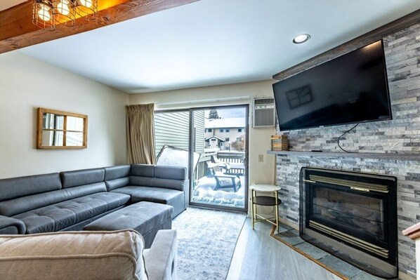 A modern living room with a sectional sofa, a flat-screen TV mounted on a stone wall, and a fireplace.
