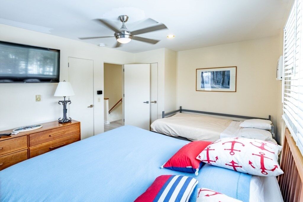 Een slaapkamer met een bed, een nachtkastje met een lamp, een plafondventilator, een lijst met een afbeelding aan de muur, en een raam met jaloezieën.