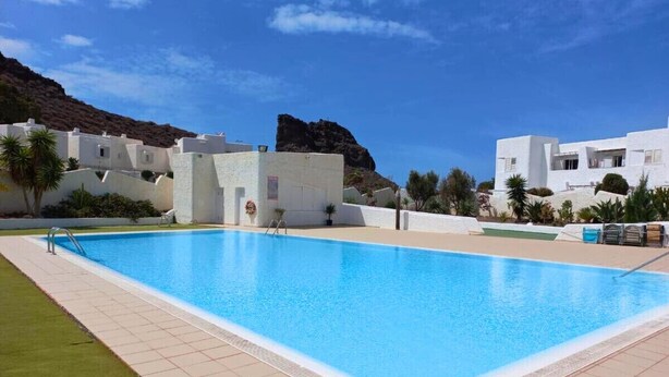 A swimming pool with a tiled deck, surrounded by white buildings and a mountainous backdrop. - 2BR Home - 3 min to Agaete Beach - Pool (Agaete)
