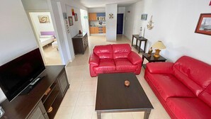A living room with a TV, red sofas, a coffee table, and a kitchen in the background. - 2BR Home - 3 min to Agaete Beach - Pool (Agaete)