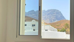 A room with a wooden dining table and chairs, a window with a view of white buildings and a mountain. - 2BR Home - 3 min to Agaete Beach - Pool (Agaete)