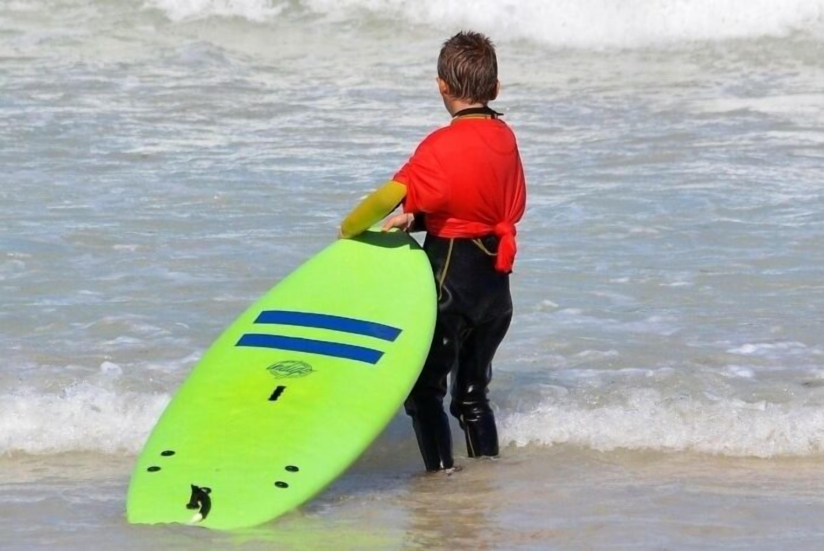 A person in a red shirt holding a green surfboard with blue stripes and a logo, standing at the edge of the water.