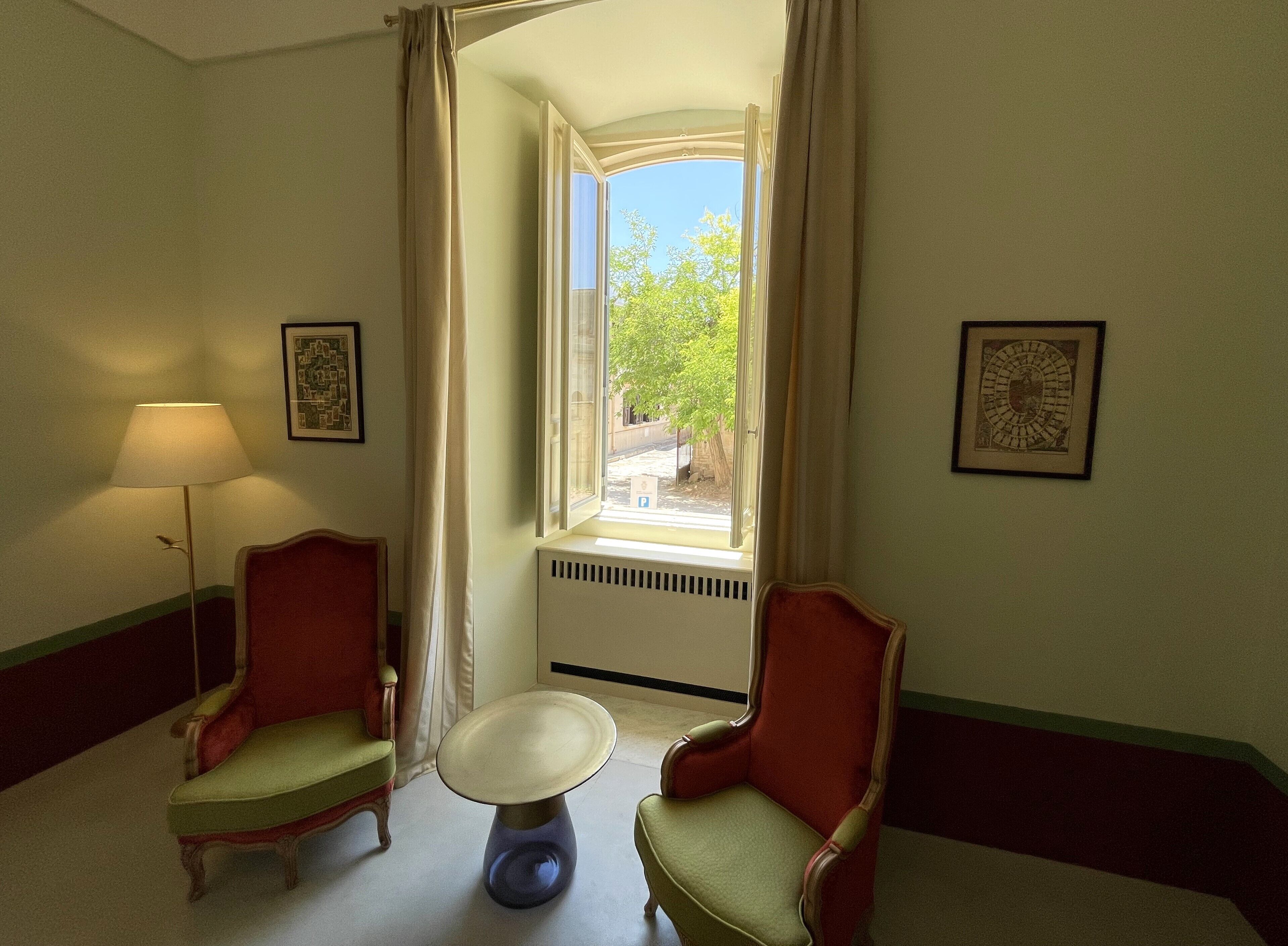 A room with two chairs, a table, a lamp, and a window with a view of greenery.