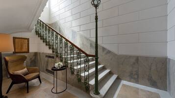 A grand staircase with marble steps and green metal railings, a plush armchair, and a small table with a vase of flowers.