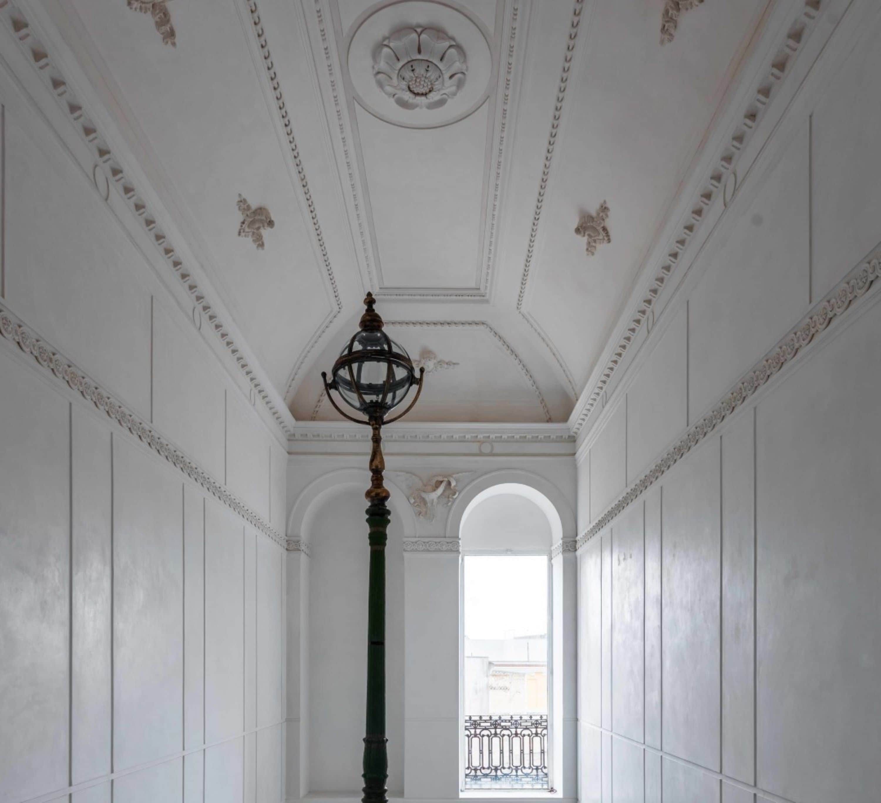 A hallway with white walls, a green column, and a ceiling with decorative moldings and a central light fixture.