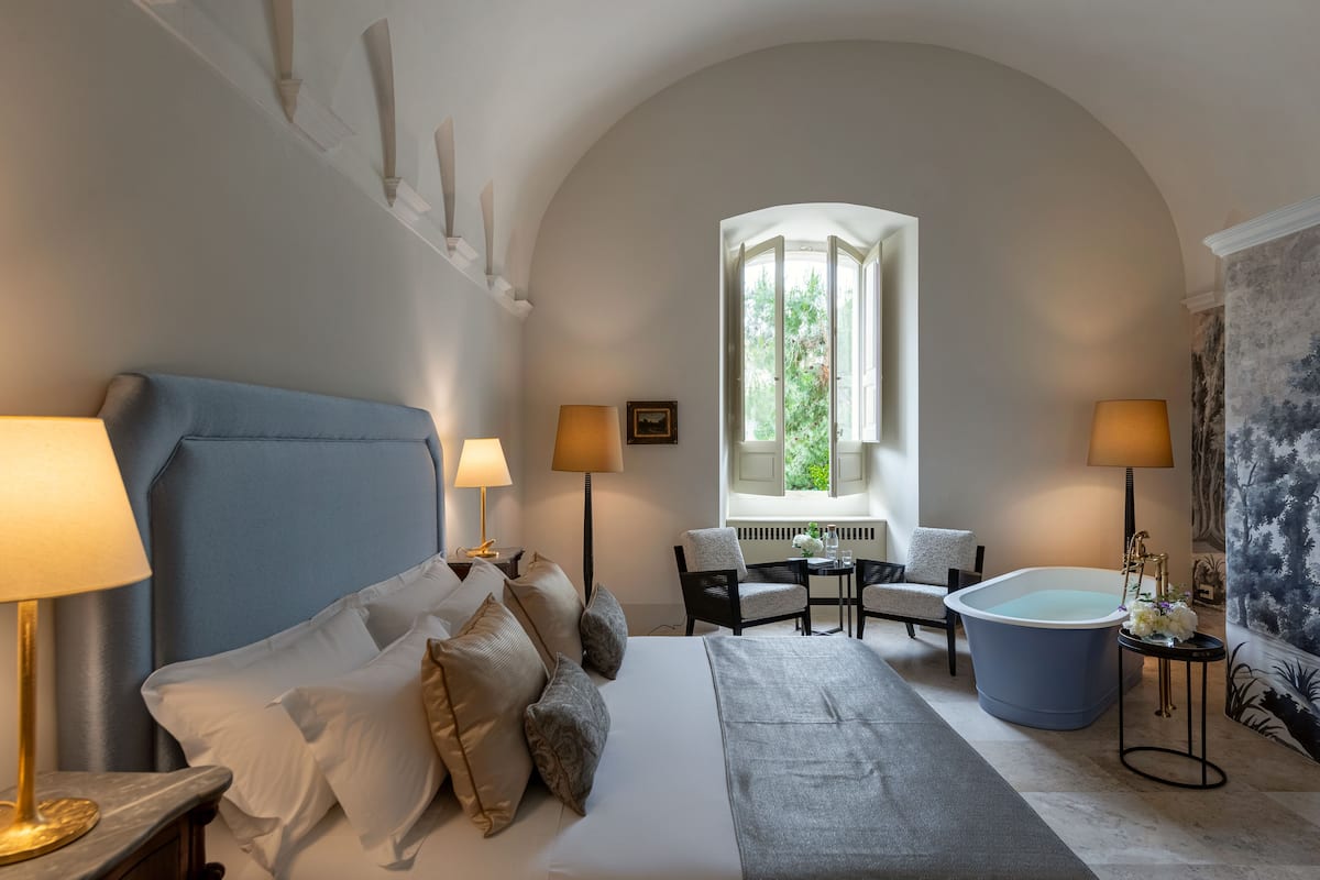 Ein Schlafzimmer mit einem großen Bett, zwei Nachttischlampen, einem Sessel, einem Tisch, einer Badewanne und einem Fenster mit Blick auf Grünflächen.