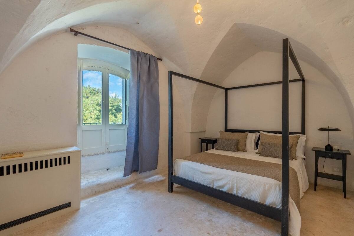 Ein Bett mit Baldachin in einem Zimmer mit schrägem Dach, einem fenster mit Vorhängen, einem Heizkörper und einem kleinen Tisch mit einer Lampe.