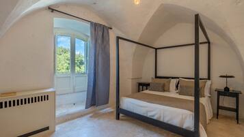 A four-poster bed in a room with a sloped ceiling, a window with curtains, a radiator, and a small table with a lamp.