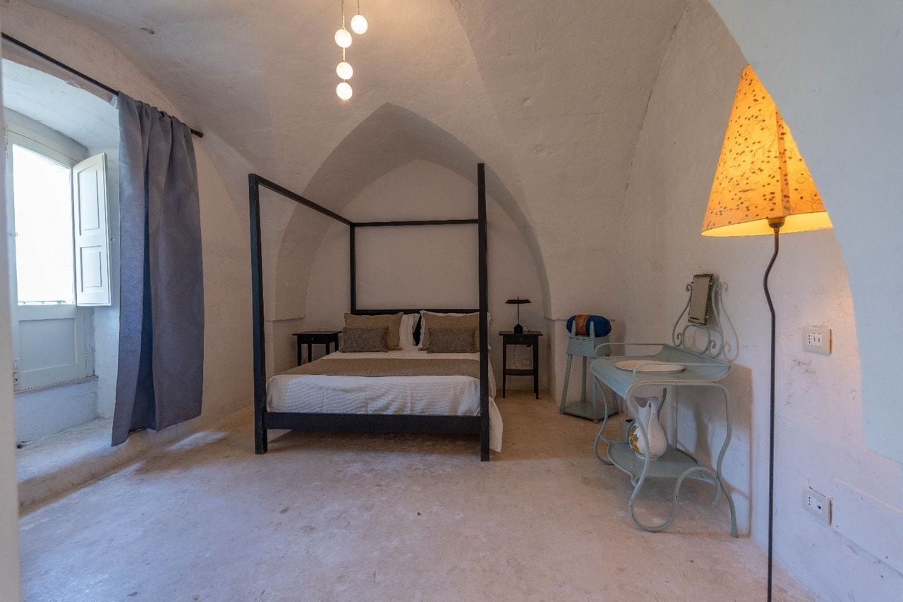 Una cama con dosel, una mesita de noche, una mesa con lámpara y una ventana con cortinas.