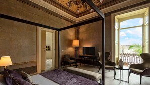 A four-poster bed with a purple bedspread, a television, a desk with a lamp, and two chairs in a room with a balcony view. - Palazzo Donna Elisabetta -Guest House (Manduria)