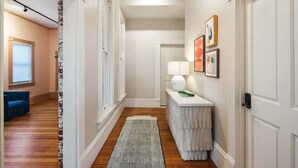 A hallway with a wooden floor, white walls, a white door, a white console table with a lamp, and framed artwork on the wall.