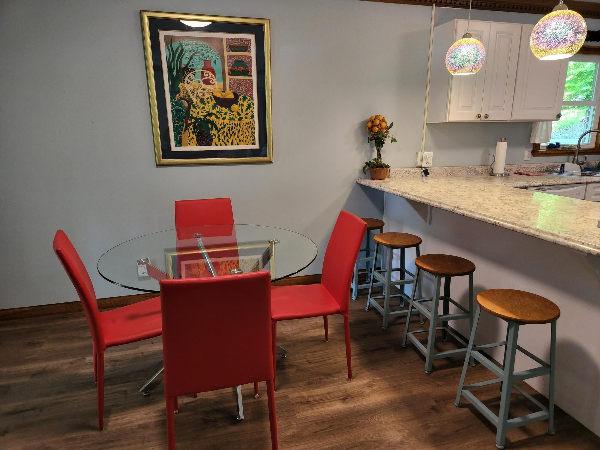 A dining area with a round glass table, red chairs, and bar stools. There is a framed artwork on the wall and a potted plant on the counter.