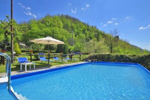Ein Swimmingpool mit blauem Wasser, umgeben von Liegestühlen und einem Tisch, mit einer Hügelkuppe und viel Grün im Hintergrund.