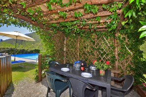 Ein Essbereich im Freien mit einem Tisch für zwei Personen, umgeben von Stühlen, einem Pool im Hintergrund und einer Holzpergola mit Kletterpflanzen.