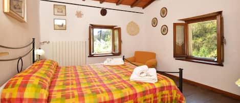 Um quarto com colcha xadrez, teto de madeira, janela com vista para vegetação e uma cadeira.