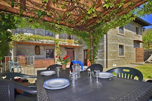 Un área de comedor al aire libre con una mesa preparada para cuatro personas, rodeada de edificios de piedra y cubierta por una pérgola de madera.