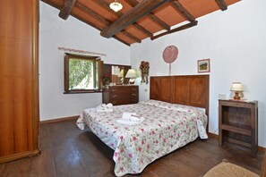 A bedroom with a wooden ceiling, a bed with a floral bedspread, a dresser, a mirror, a window, and a small table with a lamp. - Casa Le Meriche (Castiglione di Garfagnana)