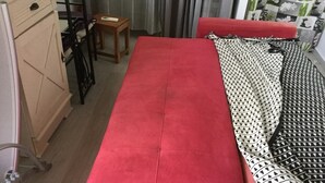 Ein Zimmer mit einem roten Schlafsofa, einem Holztisch und einer gemusterten Decke.