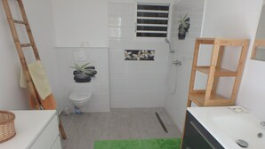 Ein Badezimmer mit Toilette, Dusche und Waschbecken. Auf dem Boden liegt ein grüner Teppich und im Duschbereich befindet sich ein Holzregal.