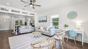 A spacious living room with a white sofa, a coffee table, and wicker chairs. A desk with a lamp and a clock is visible. - Guest Favorite Beach Home*Golf Cart*Arcade*Bikes! (Inlet Beach)