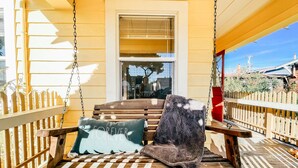 A wooden swing chair with cushions on a porch with a wooden fence and a window.