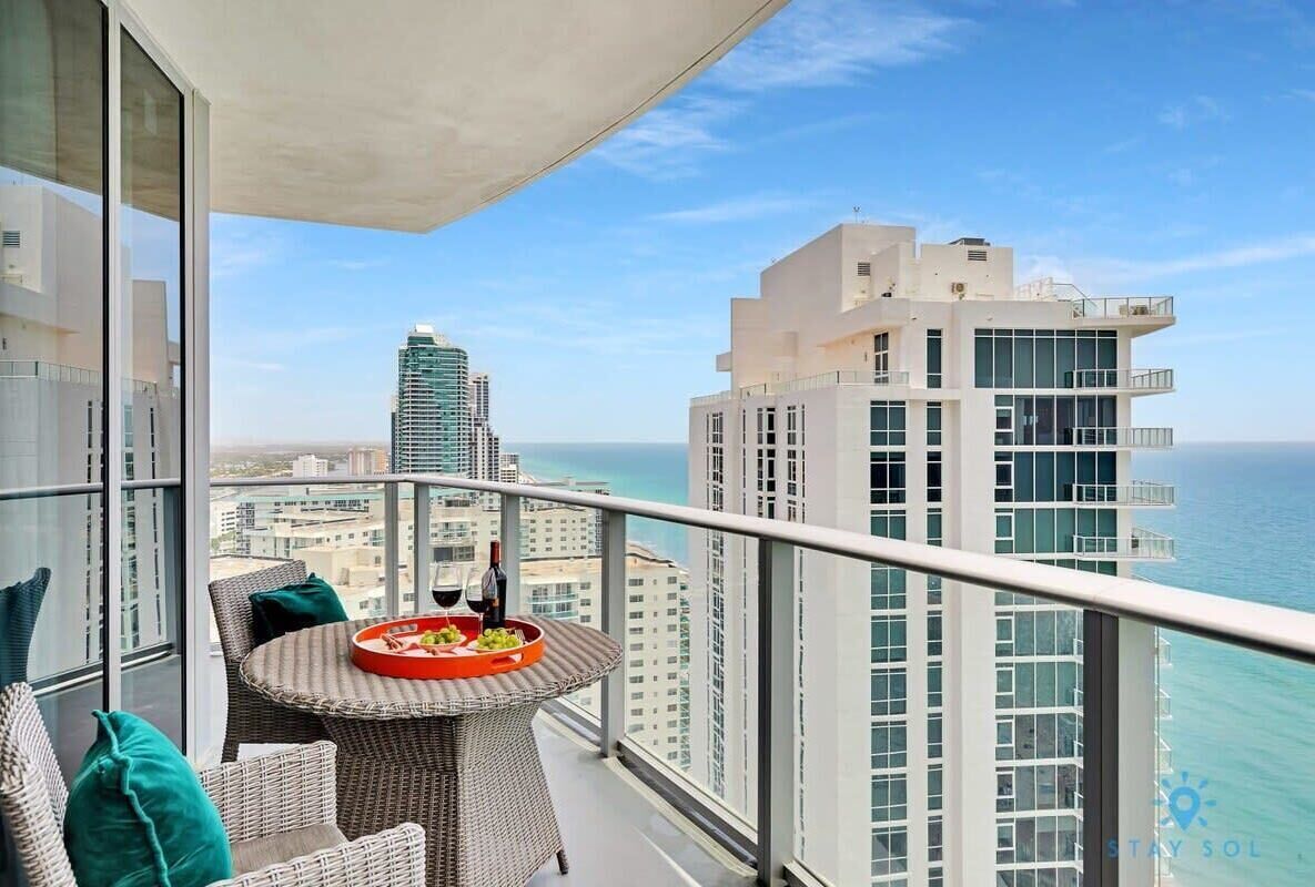 A balcony with a wicker table set for dining, overlooking a cityscape and the ocean.