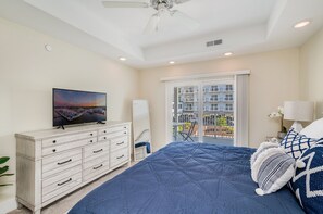 Quarto com uma cama grande, uma TV em uma cômoda, um ventilador de teto e uma varanda com vista para edifícios.