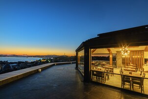 Un área de comedor al aire libre con mesas y sillas, con vista a un cuerpo de agua y el horizonte de la ciudad al anochecer.