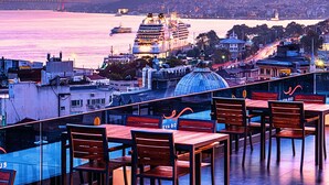 Una terraza en la azotea con mesas y sillas de madera, con vistas a un paisaje urbano que incluye una gran estructura en forma de cúpula y un crucero al fondo.